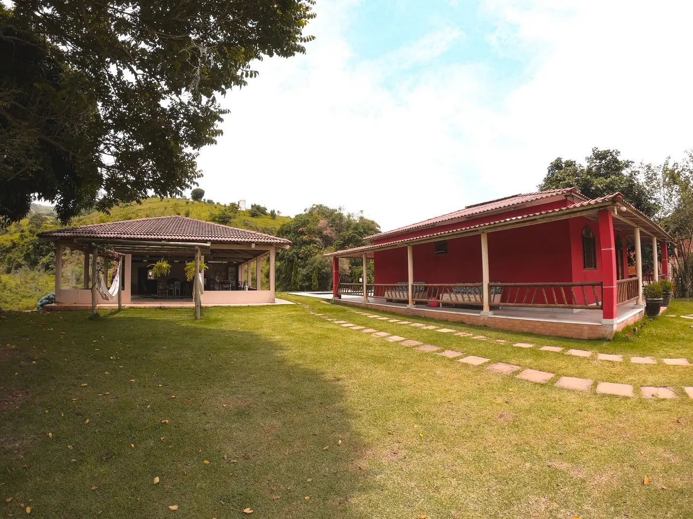 Gramado com caminho de pedras entre o salão de festas e a varanda da casa vermelha
